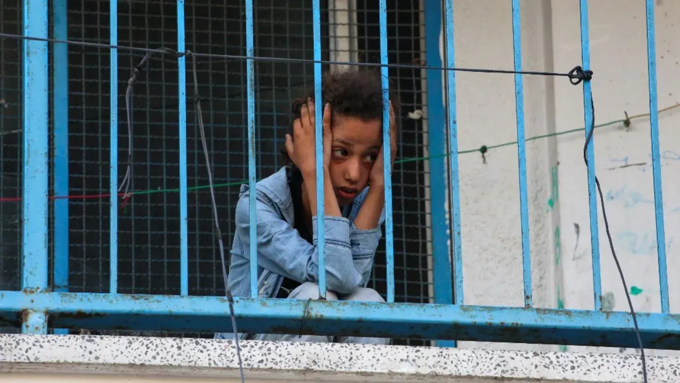 طفلة فلسطينية تجلس خلف قضبان نافذة، تعبر عن مشاعر القلق والحزن في ظل الأوضاع الإنسانية الصعبة في غزة.