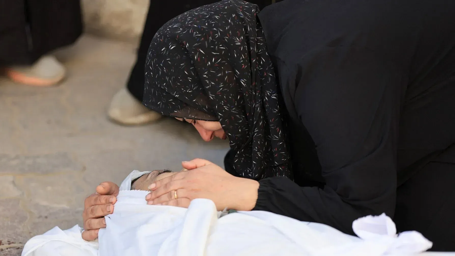امرأة ترتدي الحجاب الأسود تعانق طفلاً مغطى ببطانية بيضاء، تعبيرها يعكس الحزن والألم في ظل الظروف الصعبة في غزة.