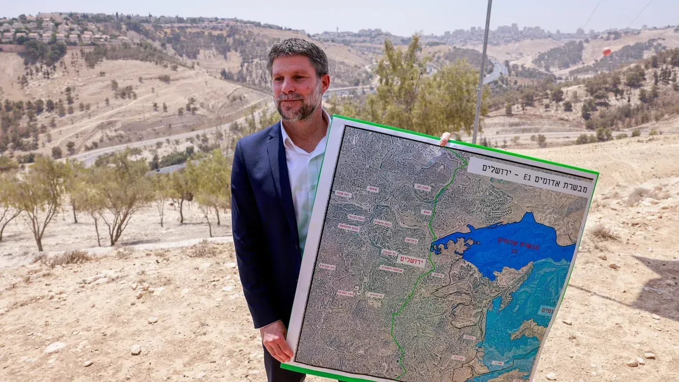 سموتريتش يحمل خريطة مشروع E1 الاستيطاني، مع خلفية للمنطقة الجبلية، في سياق خطط بناء تثير جدلاً حول مستقبل الدولة الفلسطينية.