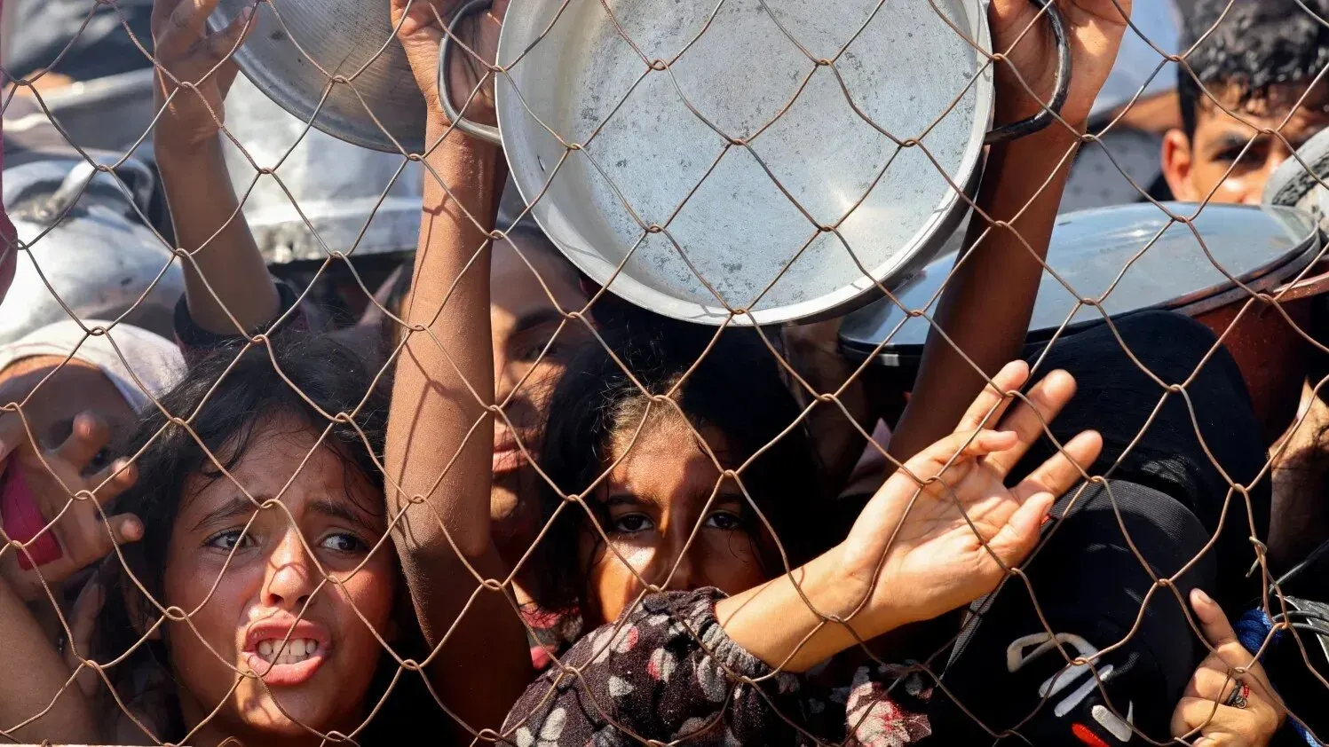 أطفال فلسطينيون يمدون أيديهم عبر سياج، يحملون أواني فارغة، بحثًا عن الطعام وسط أزمة إنسانية في غزة.