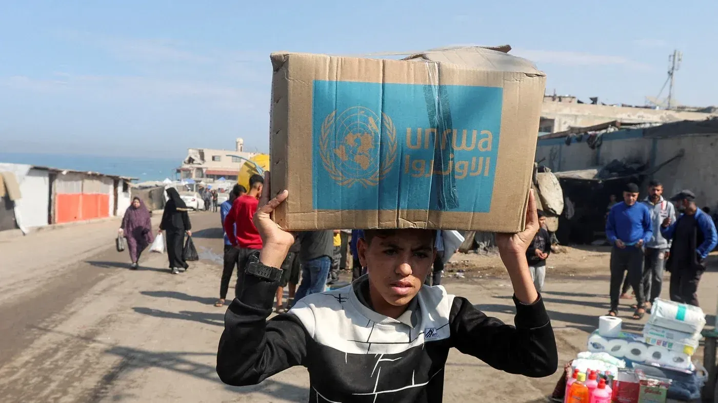 طفل يحمل صندوقًا يحمل شعار الأونروا في غزة، مع وجود آخرين في الخلفية، مما يسلط الضوء على الوضع الإنساني الصعب والمساعدات المتاحة.