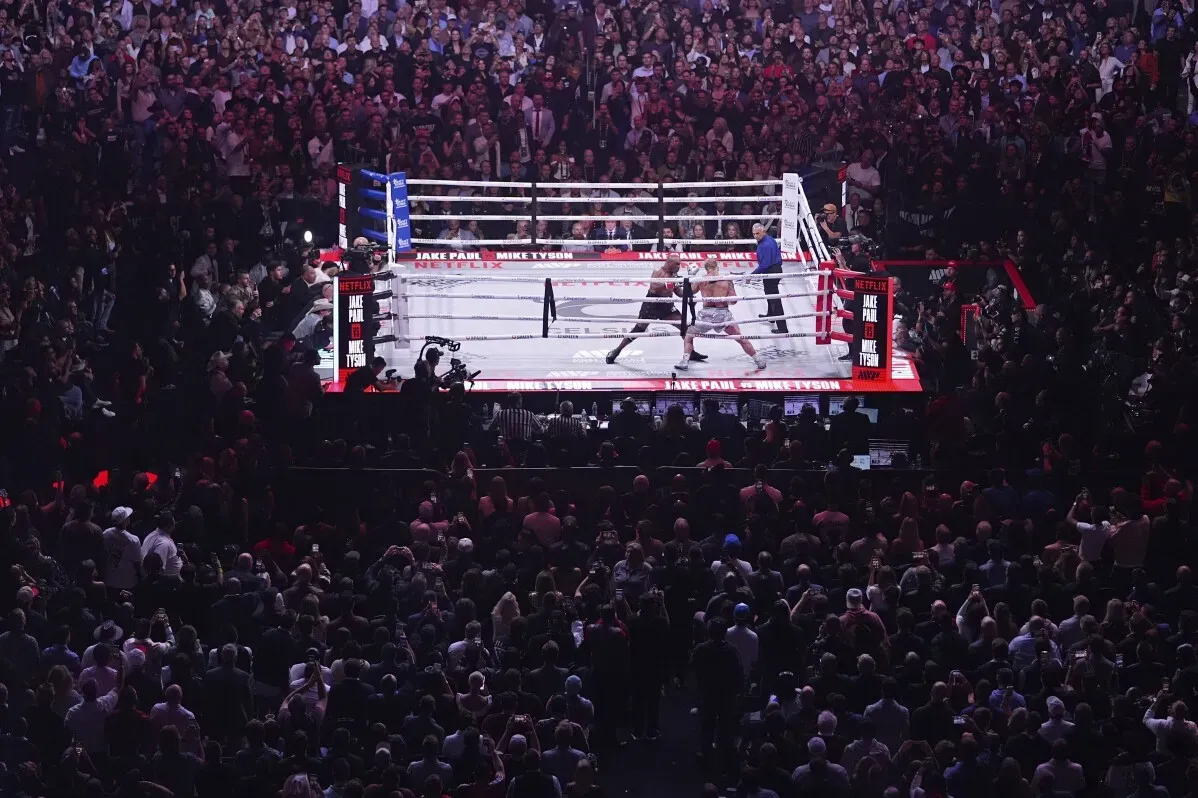 نزال مثير بين جيك بول ومايك تايسون في حلبة مملوءة بالجماهير، مع شعارات Netflix وJake Paul وMike Tyson واضحة في الصورة.