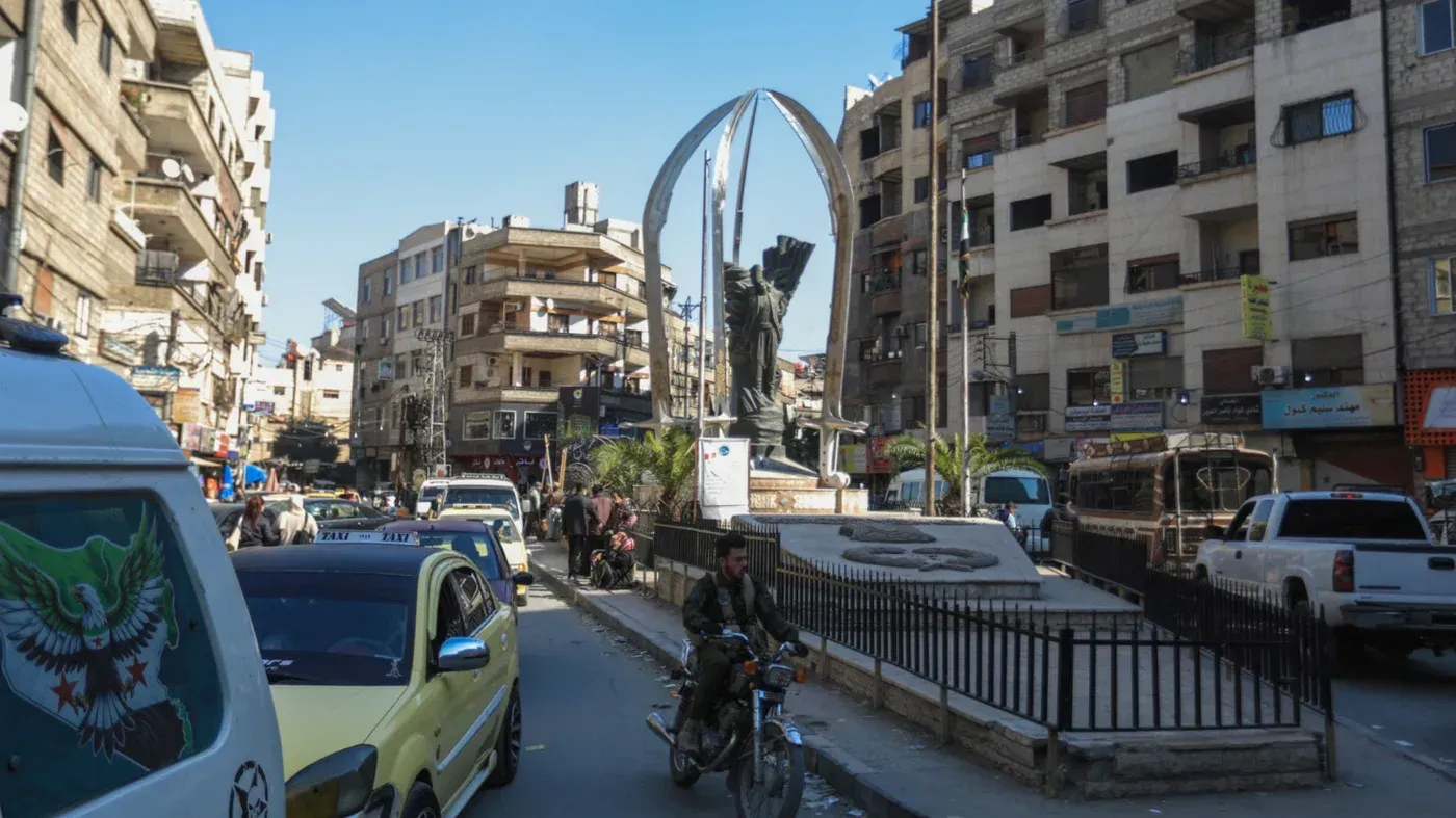 جرمانا، ضاحية جنوب شرق دمشق، تظهر تمثالًا في وسط المدينة، مع حركة مرور نشطة ومباني متعددة الطوابق، تعكس تنوع المجتمع.