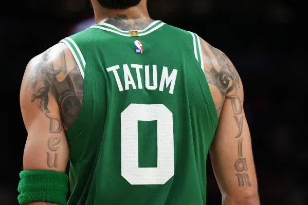 تظهر الصورة ظهر لاعب كرة السلة جيسون تاتوم مرتديًا قميصه الأخضر مع اسم "TATUM" ورقم 0، مع تاتو مرئي على ذراعه.