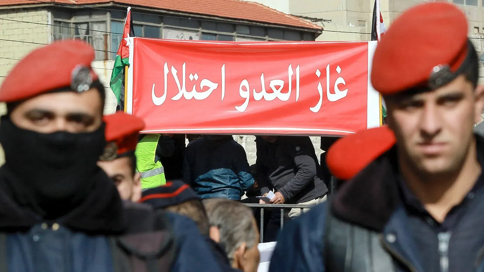 لافتة حمراء تحمل عبارة "غاز العدو احتلال" في مظاهرة أردنية، مع وجود رجال أمن يرتدون زيًا رسميًا، تعكس دعوات لإلغاء اتفاقية الغاز مع إسرائيل.