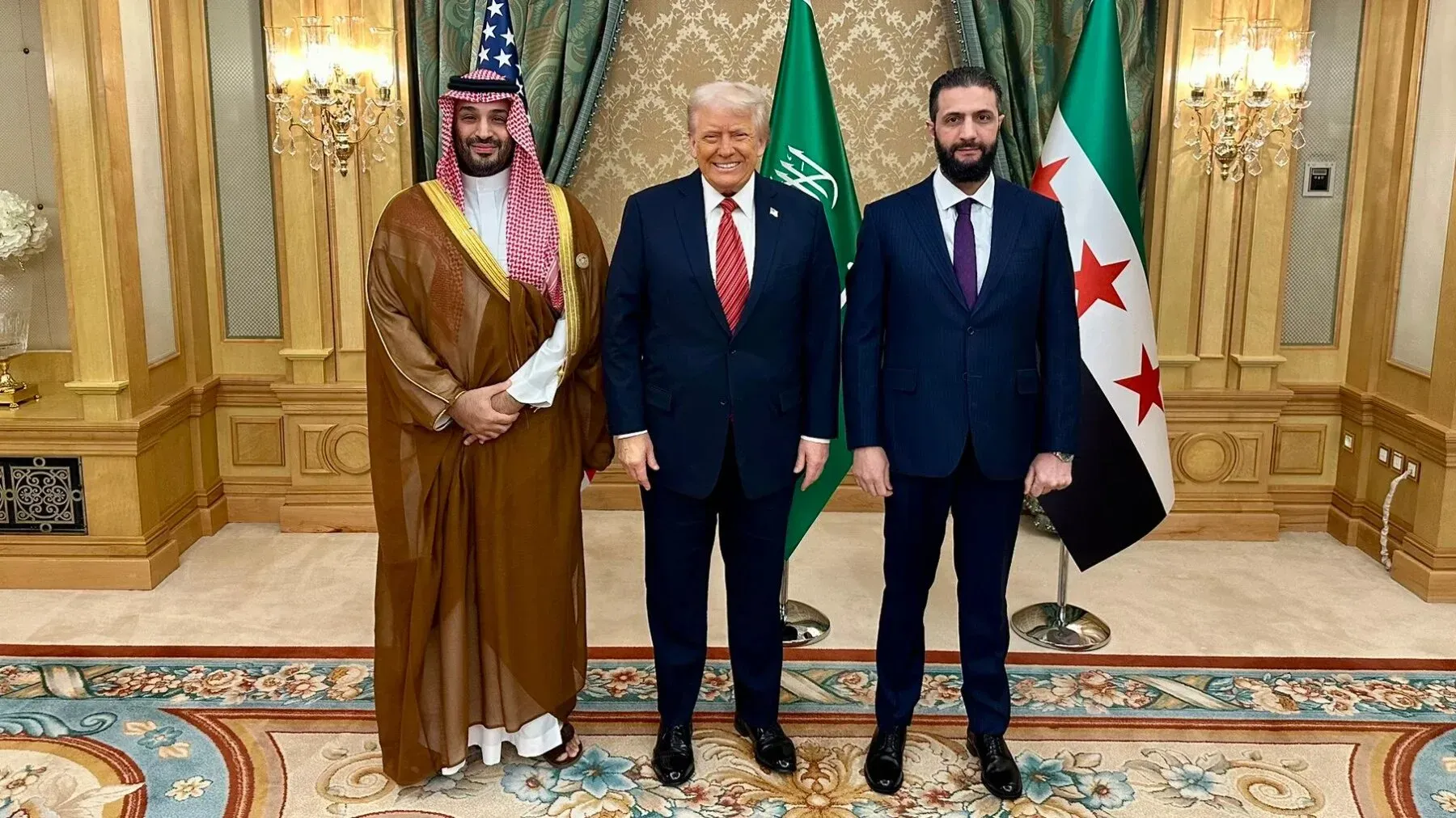 اجتماع ثلاثي بين الرئيس الأمريكي دونالد ترامب وولي العهد السعودي محمد بن سلمان والرئيس السوري أحمد الشرع، مع العلم السعودي والراية السورية في الخلفية.