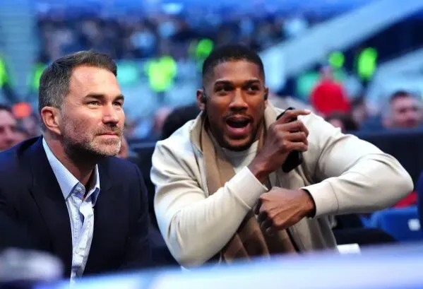 Anthony Joshua يتحدث بحماس مع مديره Eddie Hearn في حدث رياضي، مع خلفية تضم جمهورًا في الملعب.