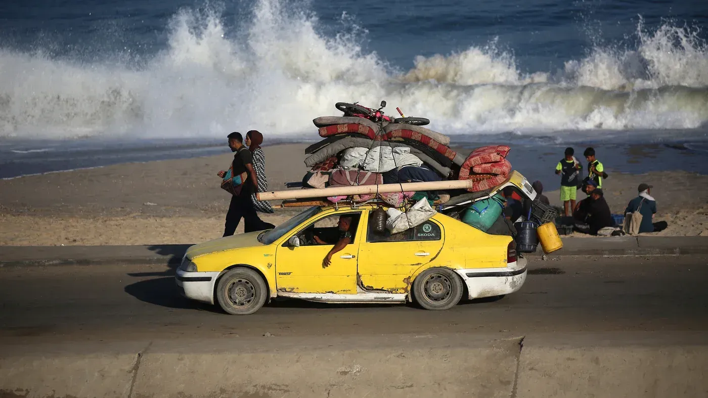 سيارة صفراء محملة بالأمتعة تسير على طريق قرب شاطئ غزة، بينما يتجمع أشخاص على الرمال، مشهد يعكس معاناة الفلسطينيين في ظل الأوضاع الصعبة.