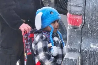 طفل يرتدي قبعة زرقاء على شكل أرنب وحقيبة ظهر، يقف بجوار سيارة في أجواء شتوية، مع ضابط هجرة خلفه.