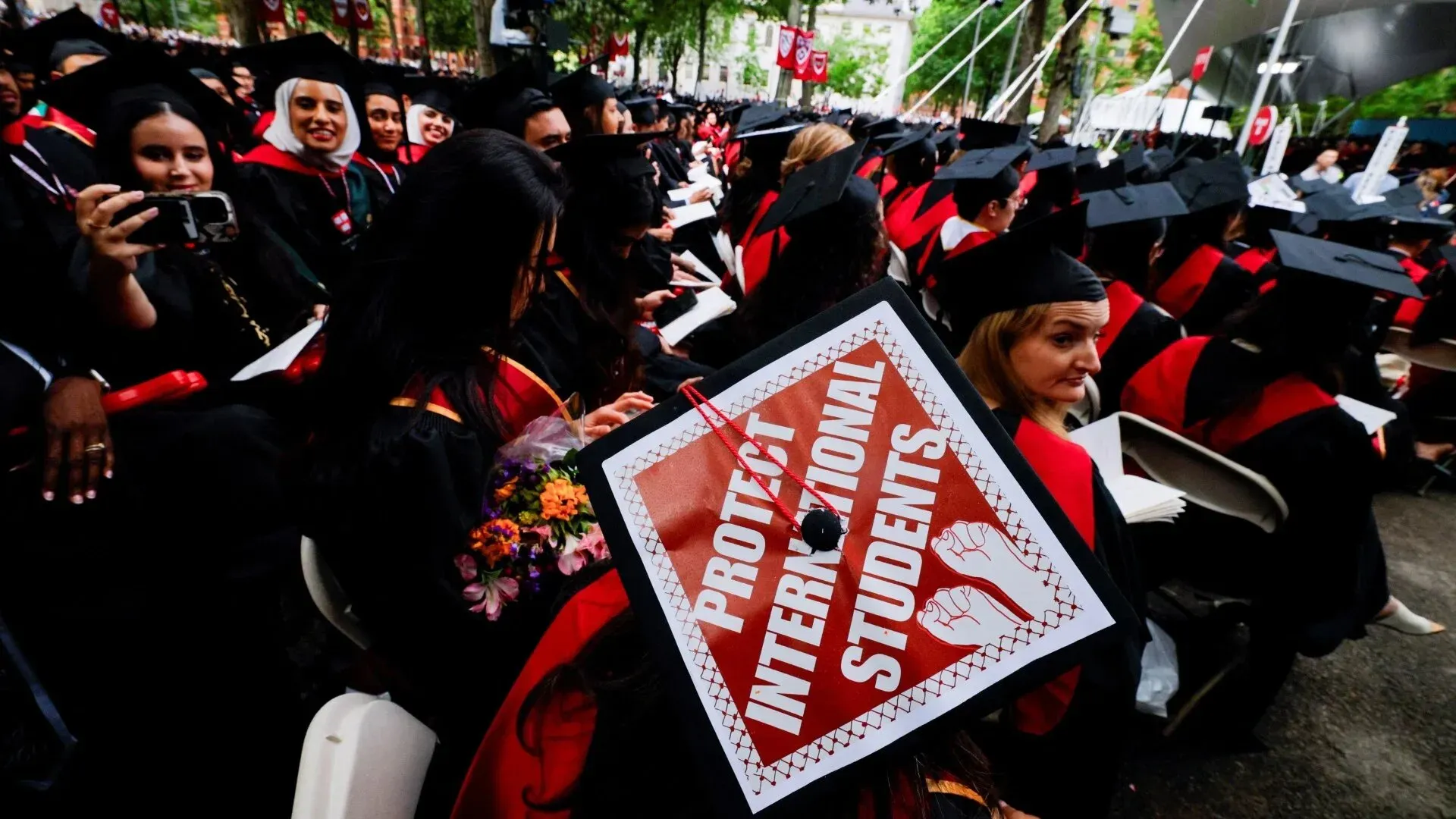 طلاب جامعة هارفارد في حفل التخرج، يحمل أحدهم لافتة تطالب بحماية الطلاب الدوليين، وسط أجواء احتفالية.
