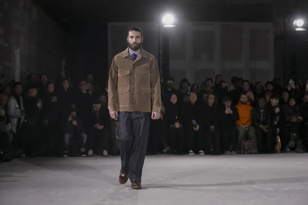 عارض أزياء يرتدي جاكيت صيد بيج بجيب كبير وسروال واسع أثناء عرض أزياء جونيا واتانابي، مع جمهور يتابع خلفه في إضاءة خافتة.