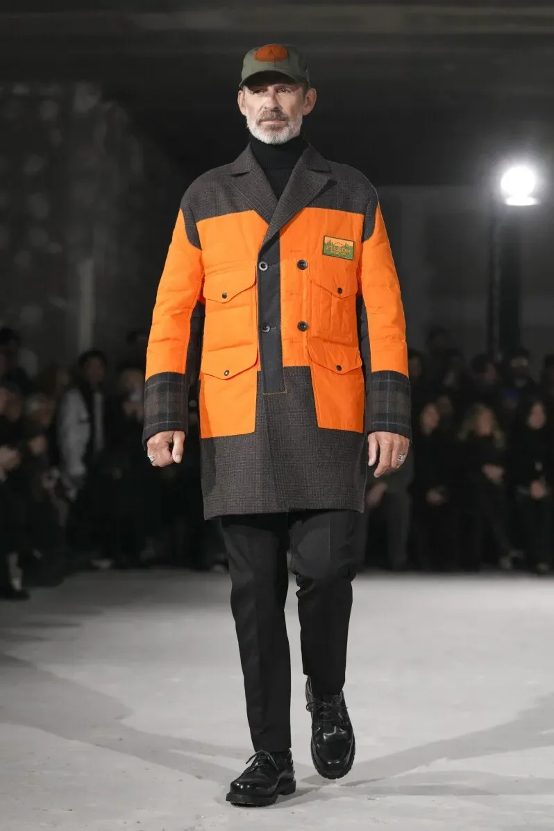 عارض أزياء يرتدي جاكيت صياد برتقالي مع تفاصيل ضخمة وجينز أسود، مقدماً لمسة معاصرة على الأزياء الرجالية في أسبوع الموضة في باريس.