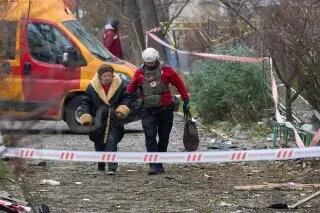 مسعف يساعد امرأة مسنّة على السير في منطقة متضررة، مع وجود مركبة طوارئ في الخلفية وحواجز أمان، مما يعكس آثار النزاع في أوكرانيا.