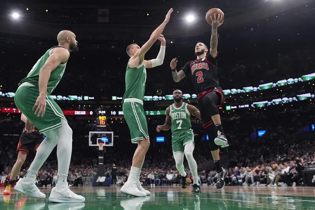 لاعب شيكاغو بولز ينفذ تسديدة أثناء تدخل لاعبين من فريق بوسطن سيلتيكس في مباراة كرة السلة، مع وجود الجمهور في الخلفية.