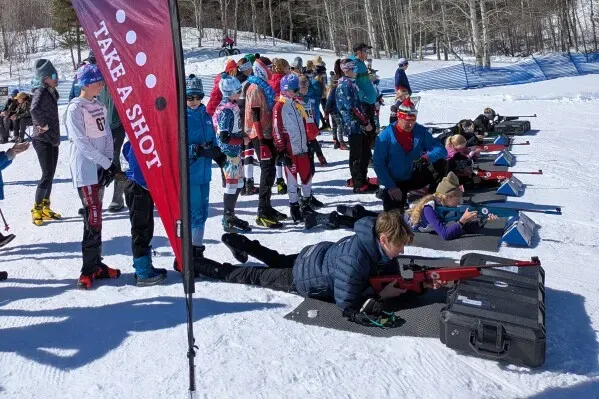 مجموعة من المتزلجين الشباب يتدربون على الرماية باستخدام بنادق الليزر في ميدان للبياثلون، مع لافتة تدعوهم لتجربة الرياضة.