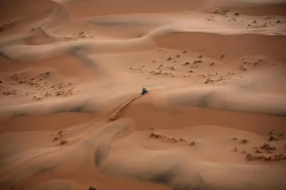 دراج يتسابق على الرمال في صحراء السعودية خلال رالي داكار، مع تضاريس رملية متموجة وخلفية طبيعية شاسعة.