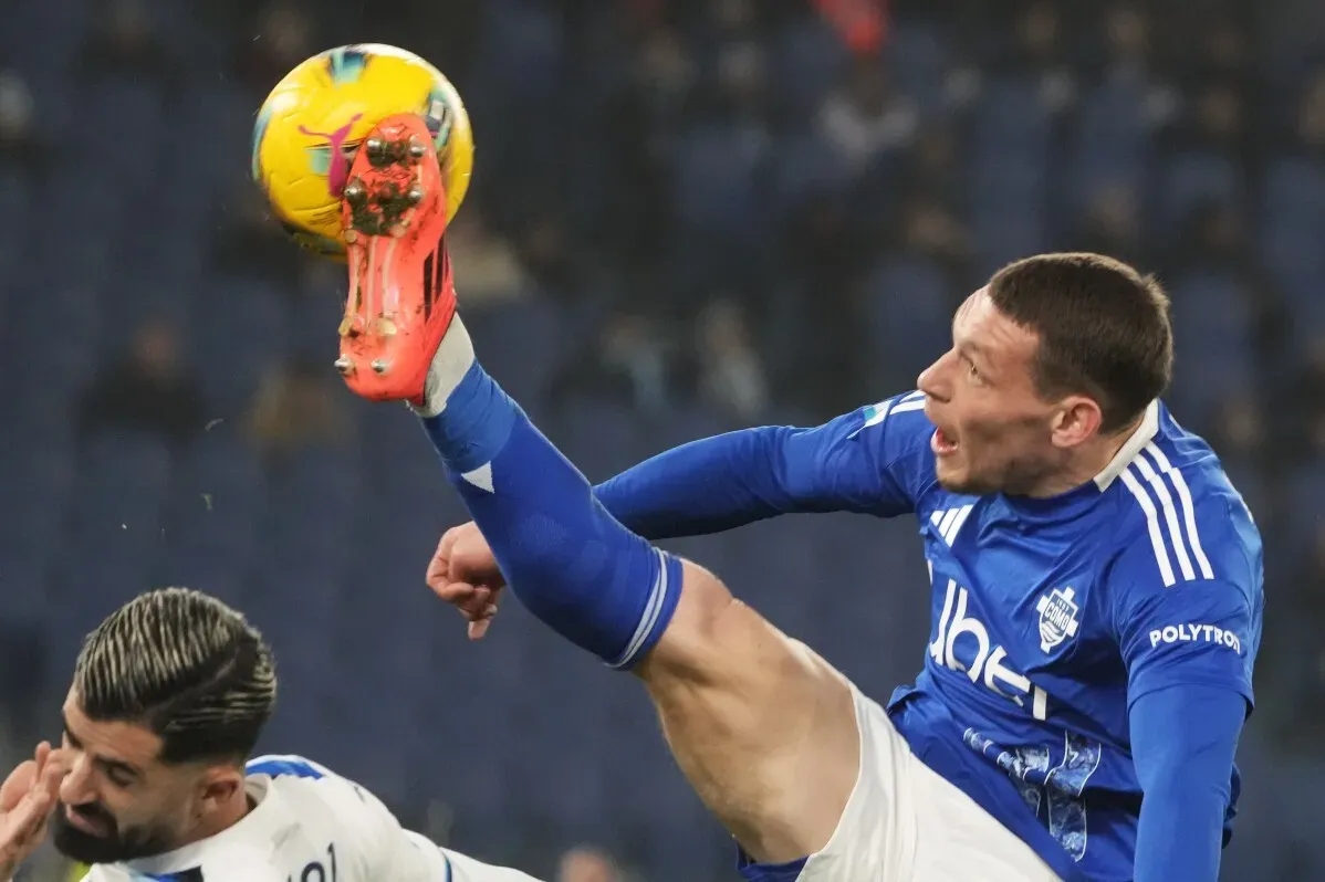 لاعب كرة قدم يرتدي زيًا أزرق، يقوم برفع قدمه عالياً لتسديد الكرة، بينما يتواجد لاعب آخر في وضعية دفاعية.