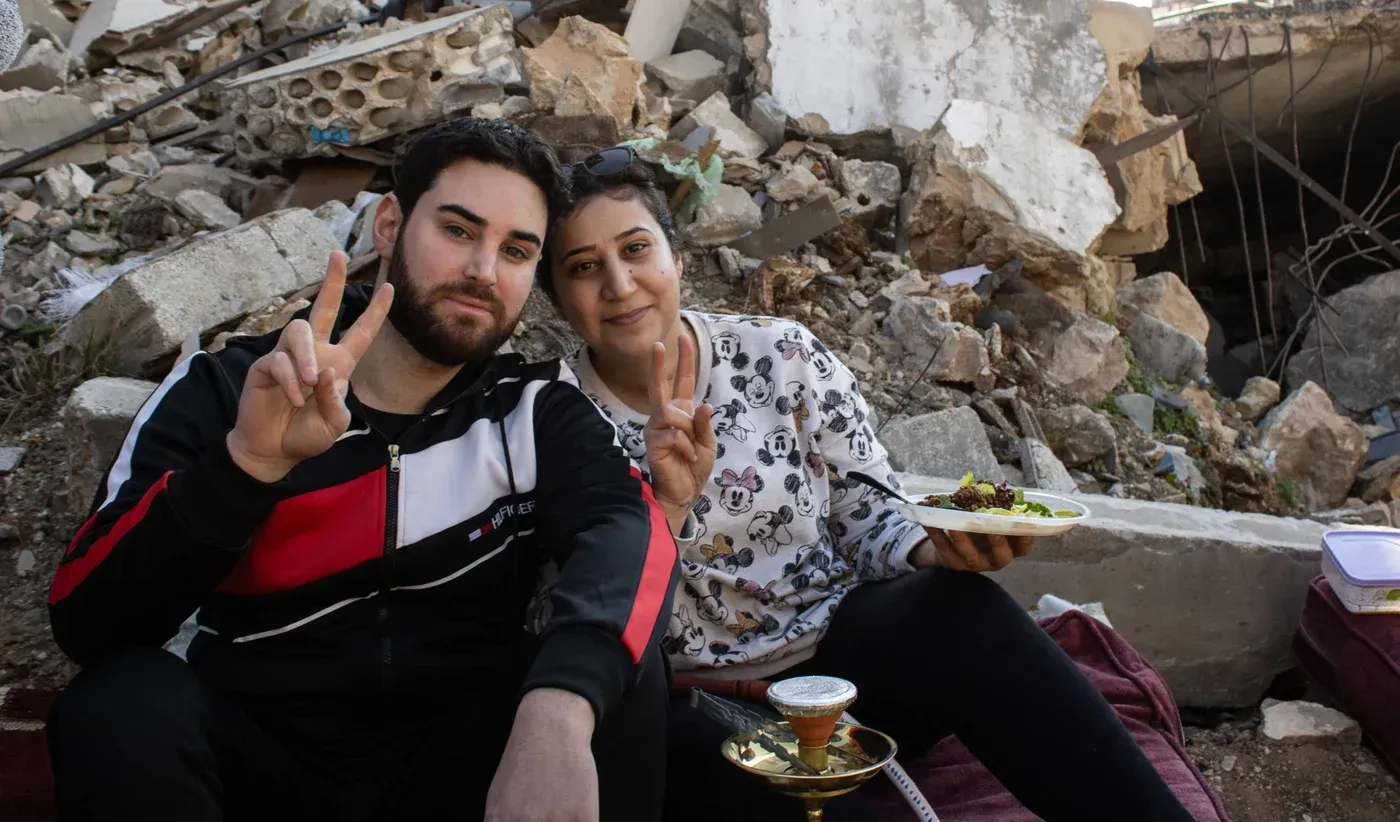 شخصان يجلسان على أنقاض مبنى في بلدة الخيام، يتبادلان الابتسامات ويحتسيان القهوة، مع وجود بقايا الحطام خلفهما.
