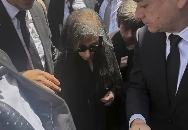 فيروز، المطربة الشهيرة، ترتدي حجابًا أسود ونظارات شمسية، محاطة بالمعزين خلال مراسم تكريم ابنها زياد الرحباني في بيروت.