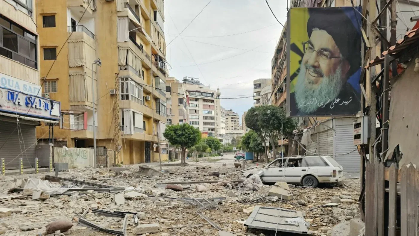 صورة لشارع مدمر في لبنان، يظهر فيه صورة كبيرة للزعيم حسن نصر الله، مع آثار الدمار والخراب في الخلفية، مما يعكس الوضع الأمني المتدهور.