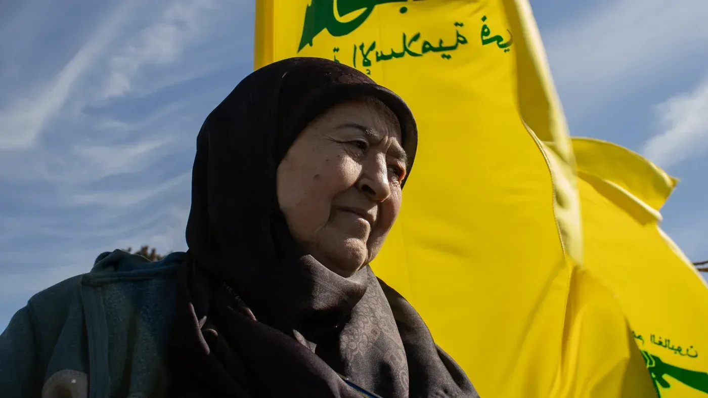 امرأة مسنّة تدعى فاطمة جعفر، تقف أمام أعلام حزب الله في قرية يارون اللبنانية، تعبر عن روح التحدي والأمل وسط التوترات الحدودية.