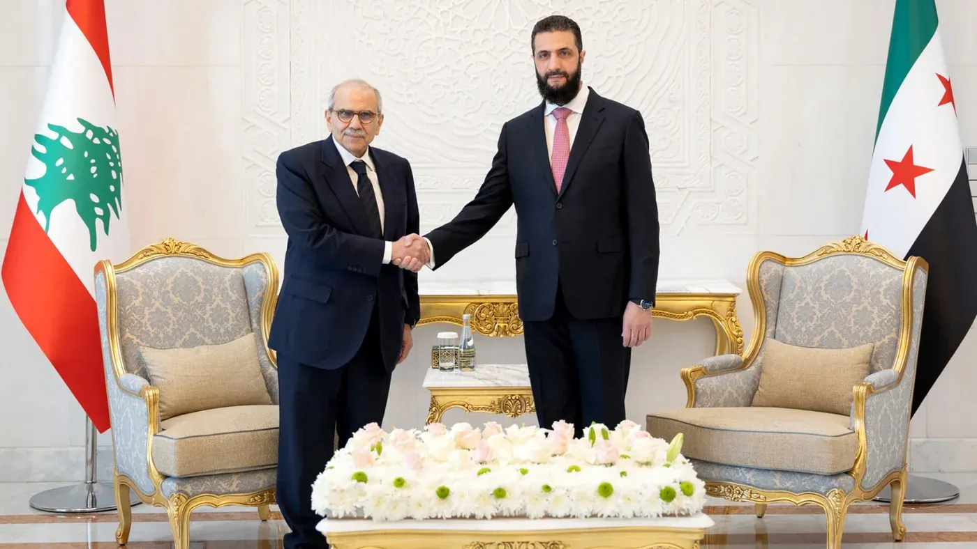 لقاء بين مسؤول لبناني وسوري، حيث يتصافحان أمام علمي لبنان وسوريا، مع تفاصيل من الأثاث الفاخر وزهور على الطاولة، يعكس التوترات السياسية بين البلدين.