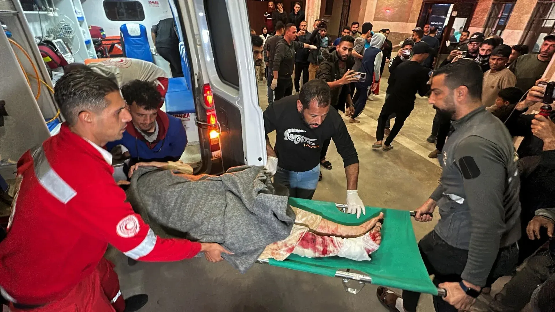 تظهر الصورة مسعفين يحملون جثمانًا ملفوفًا في بطانية، بينما يتجمع الناس حولهم في مكان مزدحم، مما يعكس الوضع الإنساني الصعب في غزة.