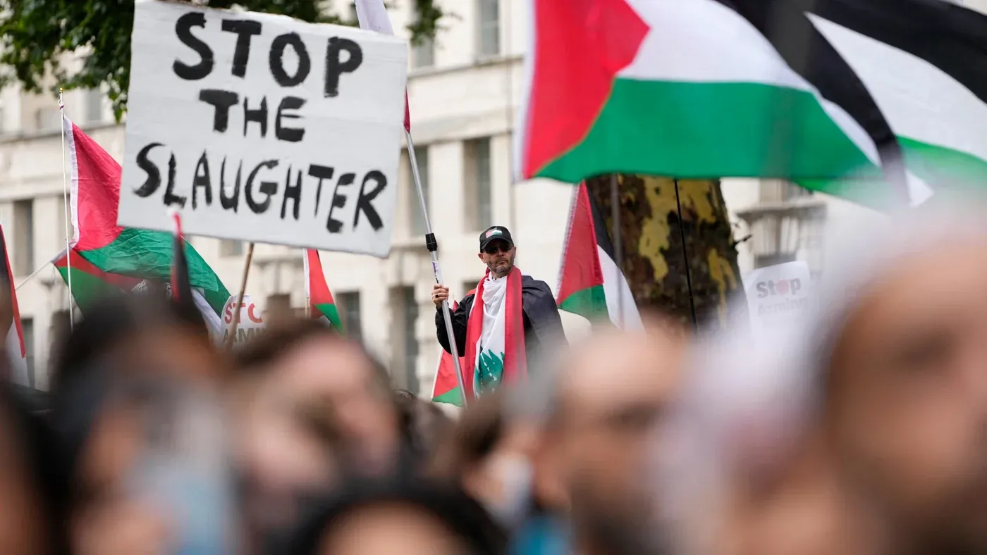 محتجون يحملون لافتة "أوقفوا المجزرة" خلال مسيرة تضامن مع فلسطين في لندن، مع أعلام فلسطينية ترفرف في الخلفية.