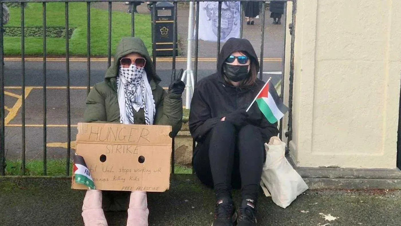 طلاب من جامعة ليستر يجلسون في إضراب عن الطعام، يحملون لافتة مكتوب عليها \"إضراب عن الطعام\" وعلم فلسطين، احتجاجًا على سياسات الجامعة.