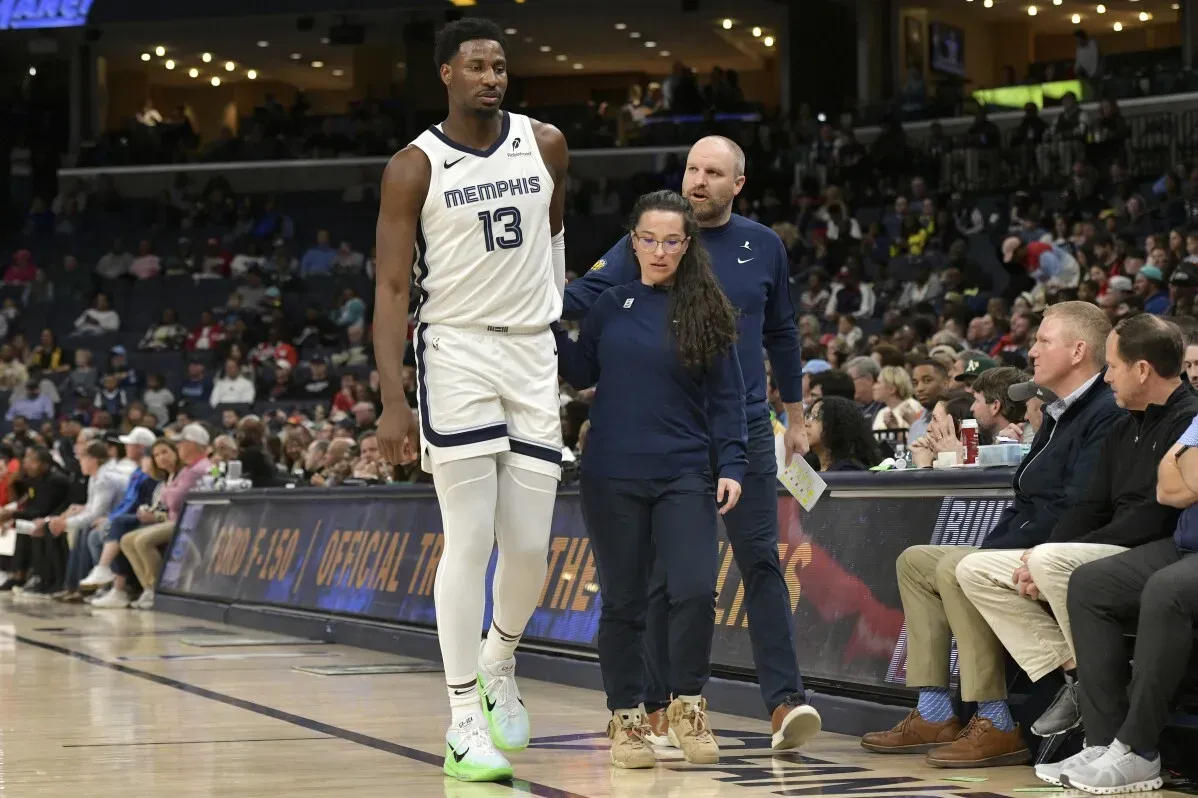 لاعب كرة سلة مصاب من فريق ممفيس جريزليز، يُساعده مدربان في الخروج من الملعب أثناء مباراة، مع جمهور في الخلفية.