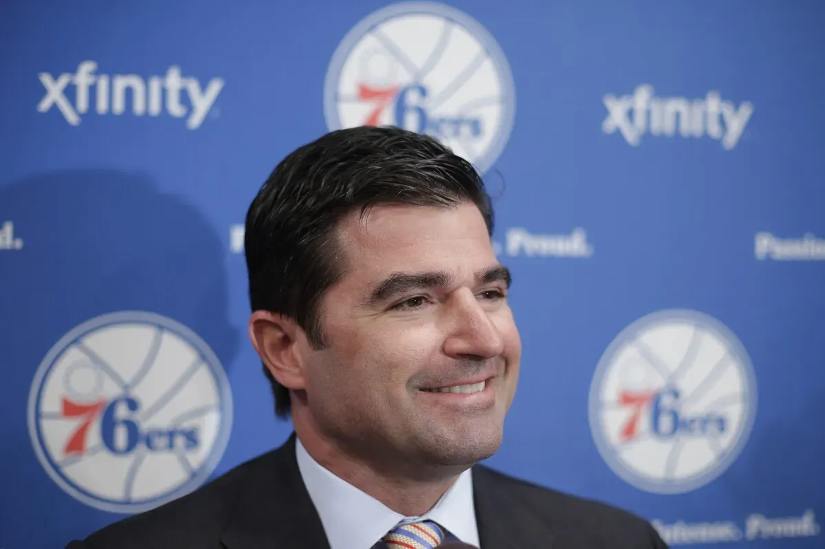 لاعب غولف مبتسم يقف أمام لافتة تحمل شعار فريق 76ers، مع خلفية زرقاء، ويعكس أجواء إيجابية في حدث رياضي.