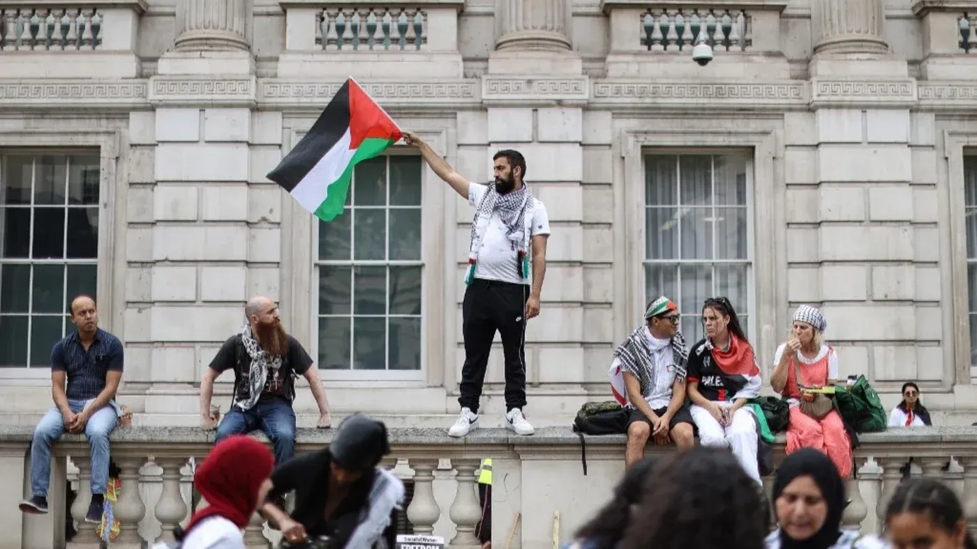متظاهر يحمل علم فلسطين في لندن، بينما يجلس آخرون على حافة مبنى، تعبيرًا عن دعمهم للقضية الفلسطينية.
