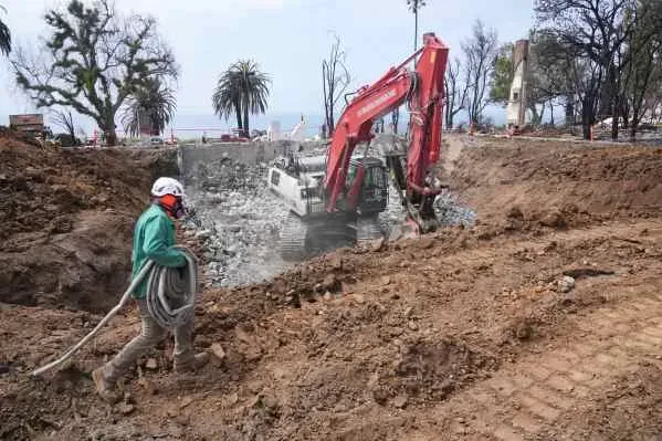عمال بناء يقومون بإزالة الأنقاض من موقع منزل دمرته حرائق الغابات في منطقة باليسادس، مع وجود جرافة في الخلفية.