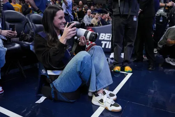 امرأة تجلس على الأرض في ملعب كرة السلة، تحمل كاميرا كبيرة وتبتسم أثناء التقاط الصور، وسط جمهور المبارة.