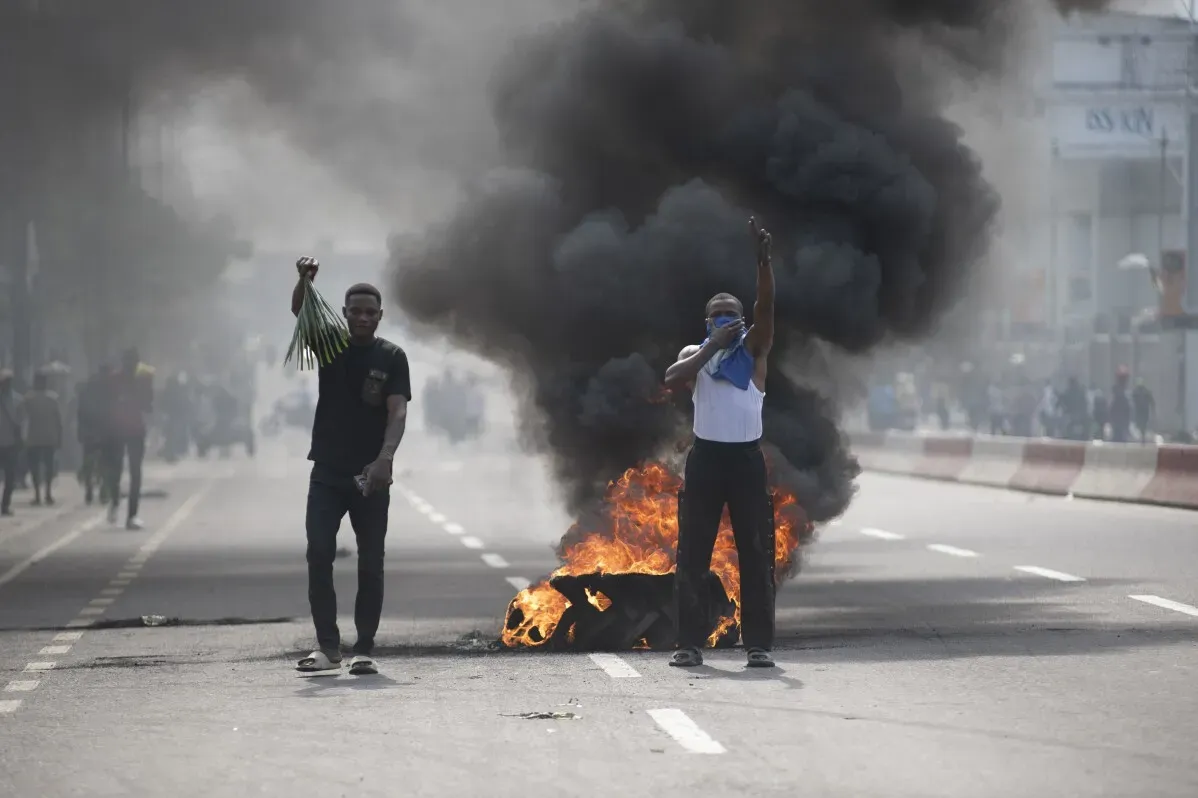 شخصان يتظاهرون في شوارع غوما مع تصاعد دخان كثيف من حريق، بينما يحمل أحدهم أغصانًا خضراء. تعكس الصورة الاضطراب الذي تشهده المدينة.