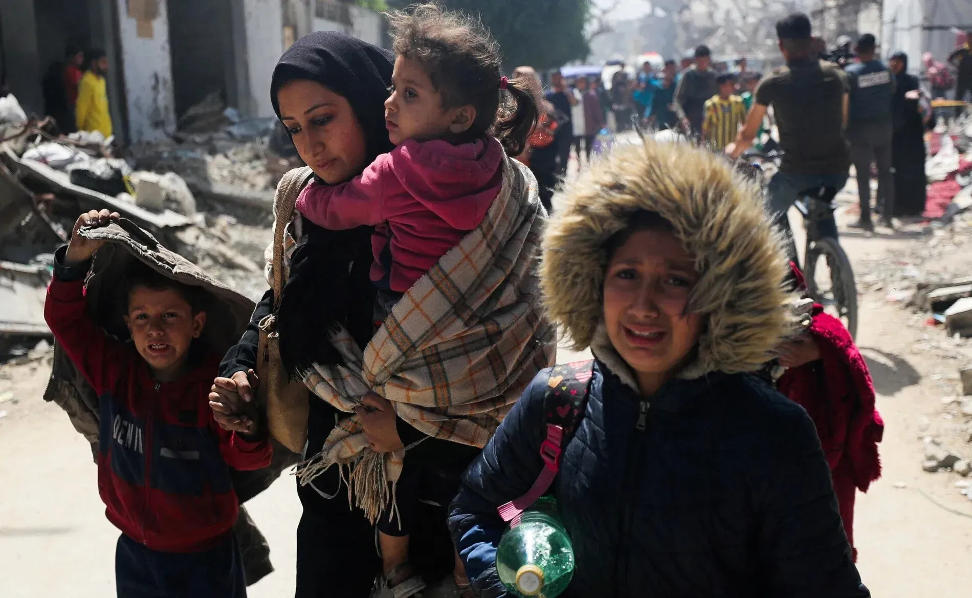 امرأة فلسطينية تحمل طفلاً على ظهرها، بينما تمسك بيدها طفلًا آخر، يسيرون في منطقة مدمرة في غزة، تعبيرًا عن معاناتهم خلال النزاع.