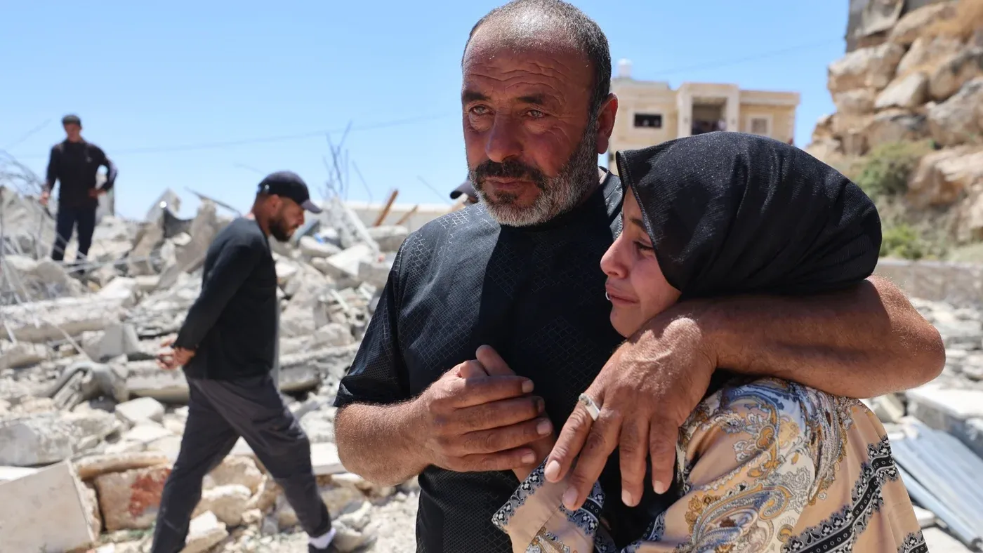 رجل فلسطيني يعانق امرأة حزينة أمام أنقاض مبانٍ مدمرة، تعبير عن الألم والمعاناة في ظل النزاع الإسرائيلي الفلسطيني.