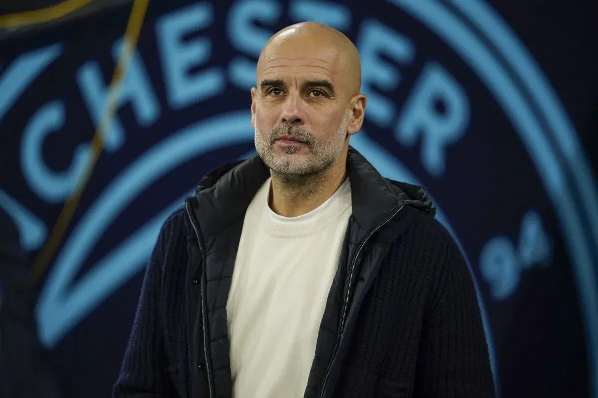 بيب جوارديولا، مدرب مانشستر سيتي، يظهر في ملعب النادي، مع خلفية شعار الفريق، بينما يناقش التحديات في الدوري الإنجليزي.