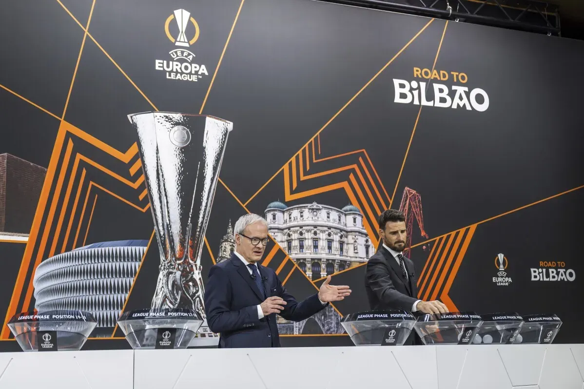 مؤتمر قرعة الدوري الأوروبي يظهر مسؤولين يتحدثان أمام خلفية تضم كأس البطولة وشعارات الأندية المشاركة.