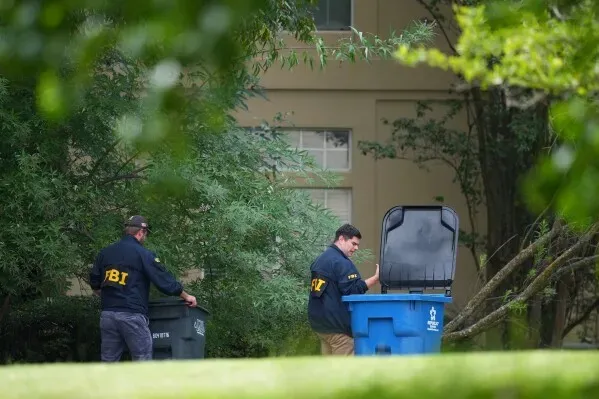 عملاء مكتب التحقيقات الفيدرالي (FBI) يقومون بتفتيش حاويات القمامة في موقع حادث إطلاق النار في دالاس، مع وجود أشجار في الخلفية.