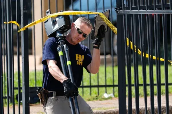 عميل من مكتب التحقيقات الفيدرالي يحمل معدات تصوير بالقرب من موقع إطلاق نار على منشأة تابعة لإدارة الهجرة والجمارك في دالاس، مع وجود شريط تحذيري في الخلفية.