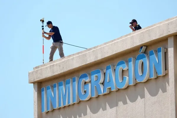 شخصان يعملان على سطح مبنى يحمل لافتة "INMIGRACIÓN"، مع معدات تصوير وأدوات، أثناء تحقيق في حادث إطلاق نار في دالاس.