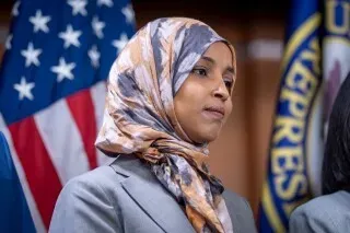 إلهان عمر تتحدث في مؤتمر صحفي، مرتدية حجابًا ملونًا، مع العلم الأمريكي خلفها، في سياق الهجوم الذي تعرضت له مؤخرًا.