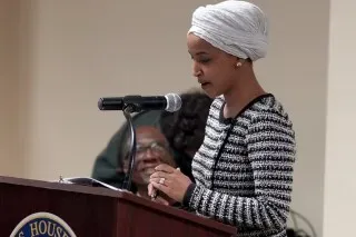 النائبة إلهان عمر تتحدث في فعالية، مرتدية زيًا رسميًا، بينما يظهر شخص آخر مبتسم خلفها، مما يعكس الأجواء السياسية في مينيابوليس.