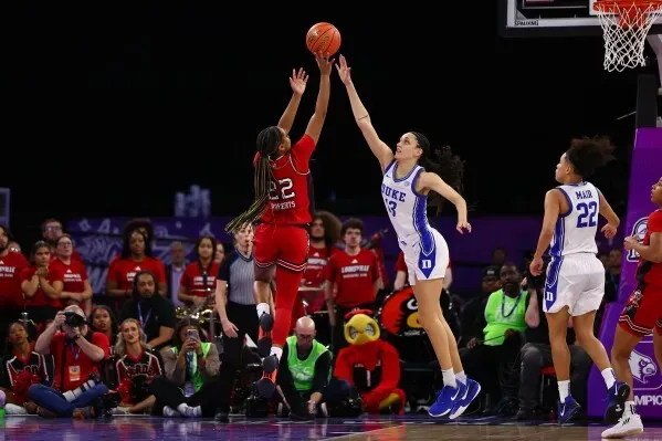 لاعبة كرة سلة من فريق لويزفيل تسدد كرة في الهواء خلال مباراة ضد ديوك، مع وجود مشجعين خلفها في المدرجات.