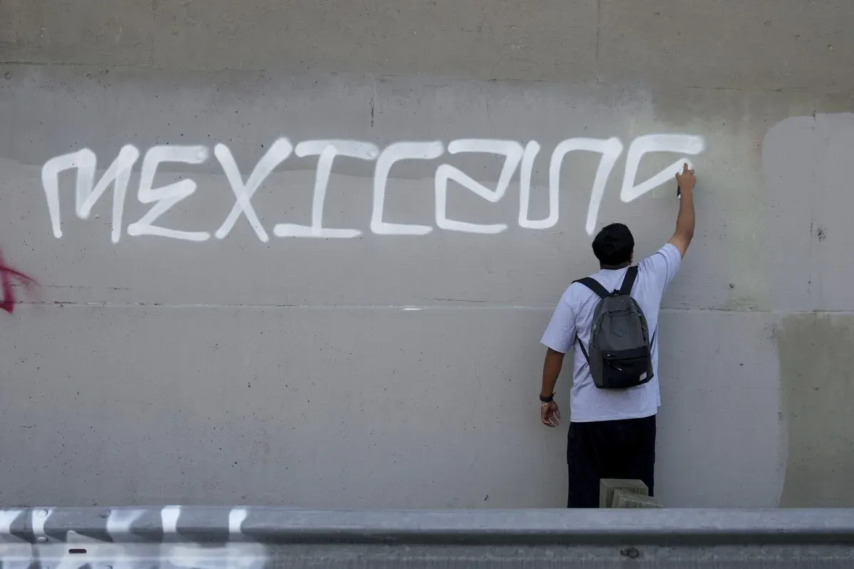 شخص يكتب كلمة "MEXICANO" باللون الأبيض على جدار رمادي، تعبيرًا عن الهوية الثقافية في سياق الاحتجاجات ضد الترحيل في لوس أنجلوس.