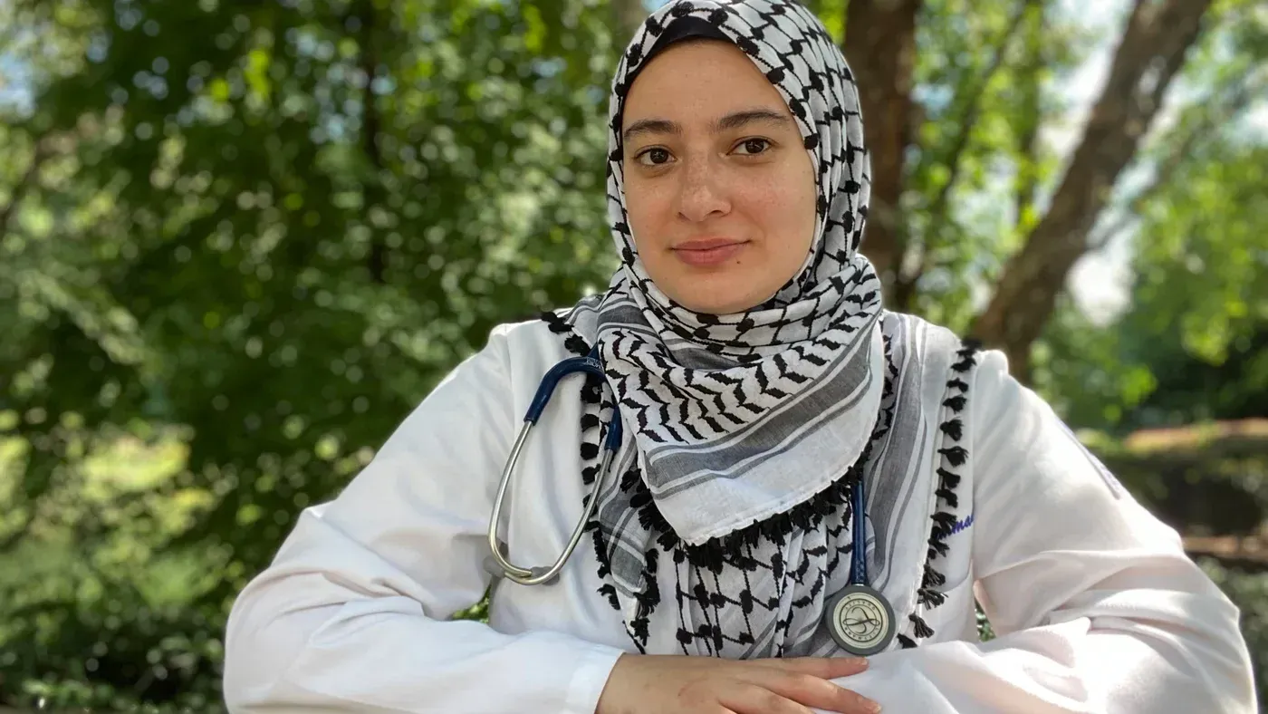 أميمة محمد، طالبة طب محجبة ترتدي معطفاً طبياً، تجلس في حديقة مع سماعة طبية حول عنقها، تعبر عن طموحاتها في مجال الرعاية الصحية.