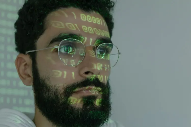 رجل ينظر بتركيز مع انعكاس أرقام ثنائية خضراء على وجهه، يعكس موضوع تحذيرات قادة الذكاء الاصطناعي من مخاطر أسراب الذكاء الاصطناعي والسيطرة على الإنترنت.