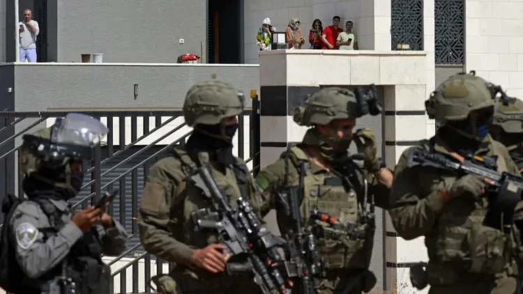 جنود إسرائيليون مدججون بالسلاح يقفون أمام مبنى في الضفة الغربية بينما يراقبهم فلسطينيون من شرفة، في سياق تصاعد العنف الاستيطاني في المنطقة.