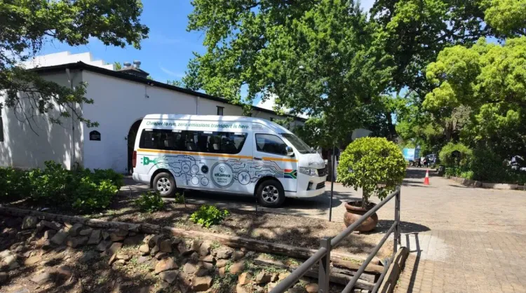 حافلة كهربائية بيضاء تحمل شعار eKamva Electric متوقفة وسط شارع مظلل بالأشجار في ستيلينبوش، تعكس توجهات التنقل الكهربائي المستدام في جنوب أفريقيا.